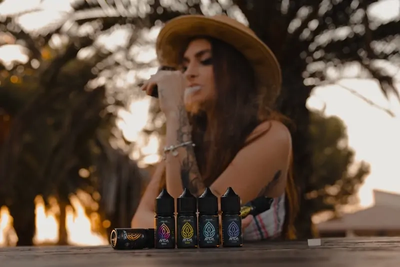 A woman taking a puff from a vape pen and exhaling. 4 bottles of Avida CBD Vape juice set in the table in front of her. 