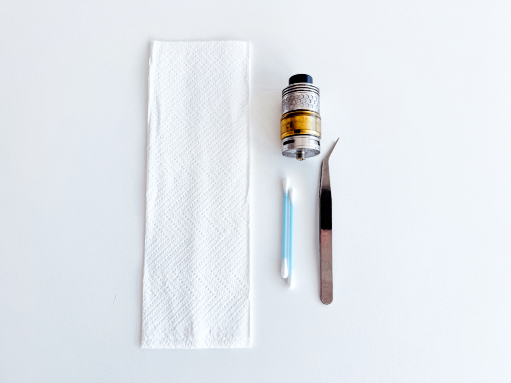 Vape cleaning kit laid out on a white surface, featuring a paper towel, cotton swab, tweezers, and a vape tank. These tools are used for maintaining vape pens and preventing clogs by cleaning residue from the tank and internal components.