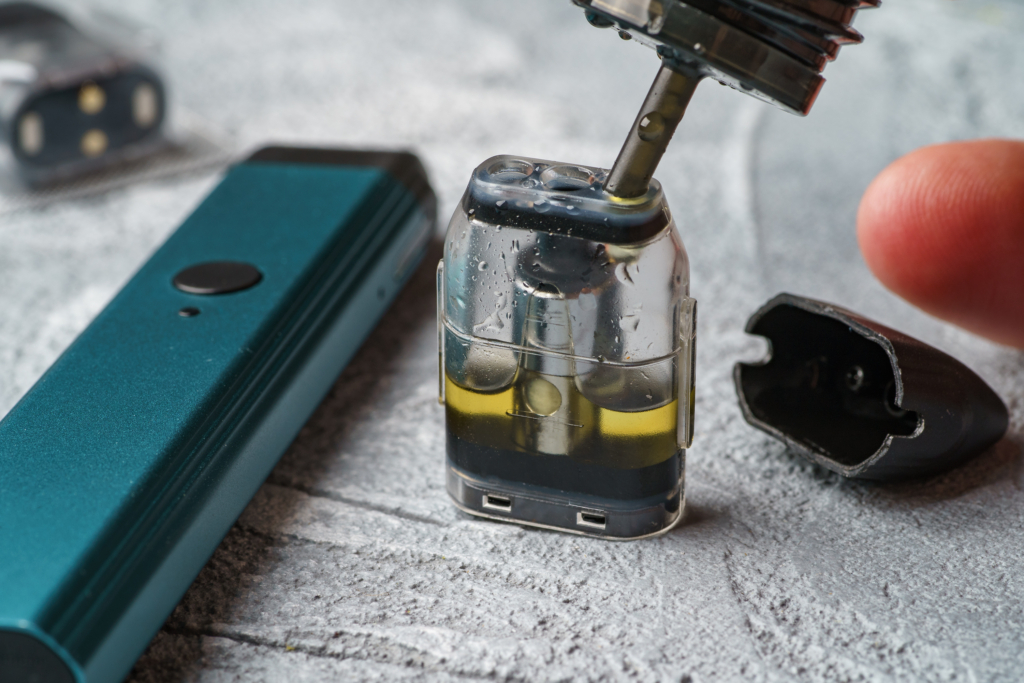 Close-up of a person refilling a vape pod with oil, showing a clear pod partially filled with e-liquid and a vape pen beside it. The pod is open, highlighting the internal components, which are often prone to clogging if not properly maintained.