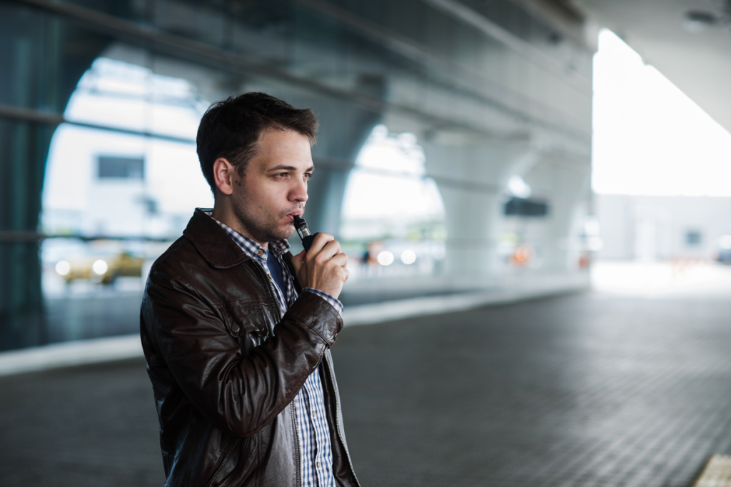 Man with hair and black jacket standing outside airport terminal vaping.