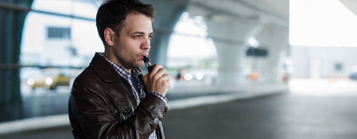 Man with hair and black jacket standing outside airport terminal vaping.